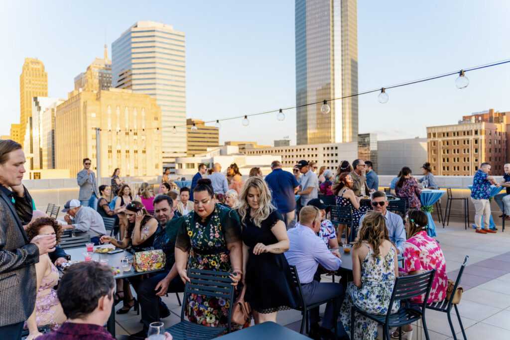 Roof terrace with a large crowd sitting at tables, enjoying food and the beautiful golden hour over downtown Oklahoma City's skyline.