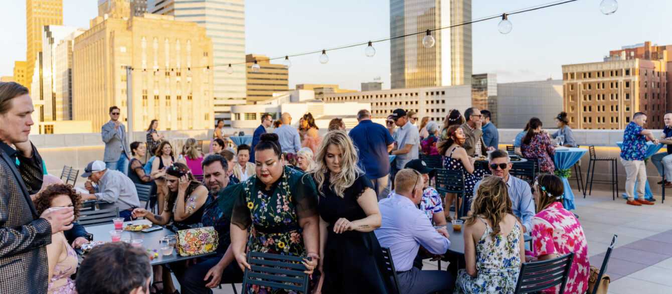 Roof terrace with a large crowd sitting at tables, enjoying food and the beautiful golden hour over downtown Oklahoma City's skyline.