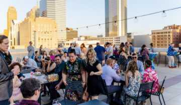 Roof terrace with a large crowd sitting at tables, enjoying food and the beautiful golden hour over downtown Oklahoma City's skyline.