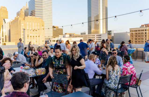 Roof terrace with a large crowd sitting at tables, enjoying food and the beautiful golden hour over downtown Oklahoma City's skyline.