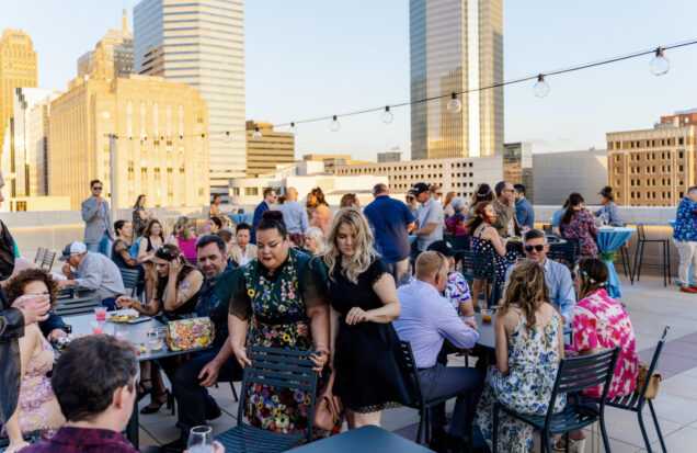 Roof terrace with a large crowd sitting at tables, enjoying food and the beautiful golden hour over downtown Oklahoma City's skyline.