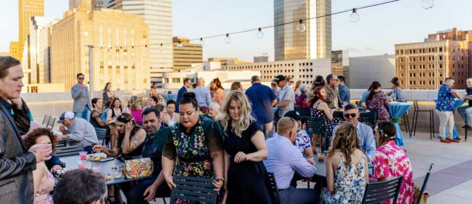 Roof terrace with a large crowd sitting at tables, enjoying food and the beautiful golden hour over downtown Oklahoma City's skyline.