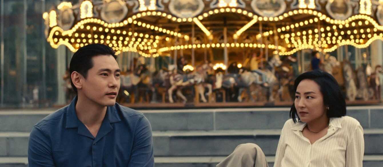 A film still from Past Lives with a man and a woman sitting next to each other in front of a carousel.