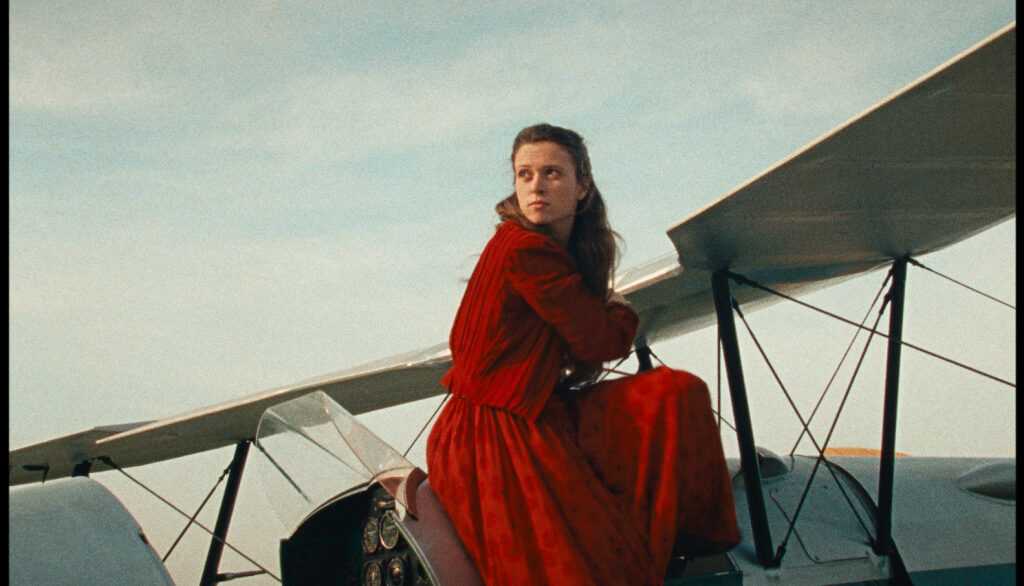 A Film Still from Scarlet with a young woman in a red dress sitting on a vintage airplane.