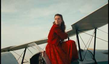 A Film Still from Scarlet with a young woman in a red dress sitting on a vintage airplane.