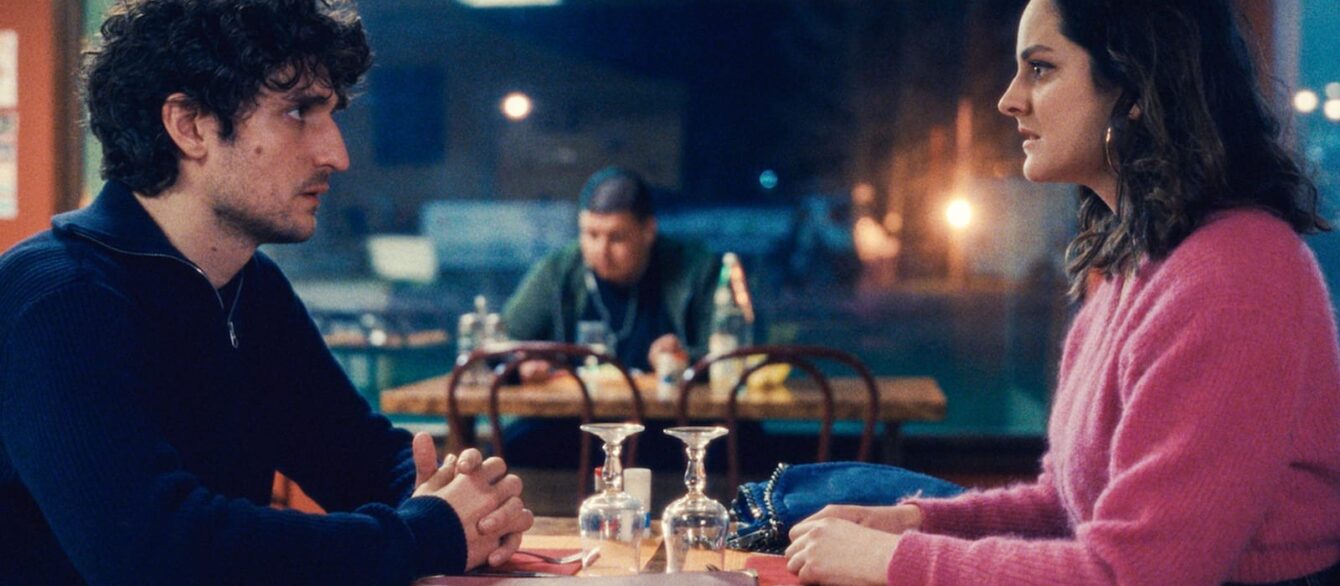 A film still from The Innocent with Louis Garrel and Noémie Merlant sitting accross from each other at a table in a cafe.