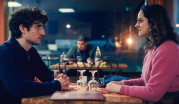 A film still from The Innocent with Louis Garrel and Noémie Merlant sitting accross from each other at a table in a cafe.