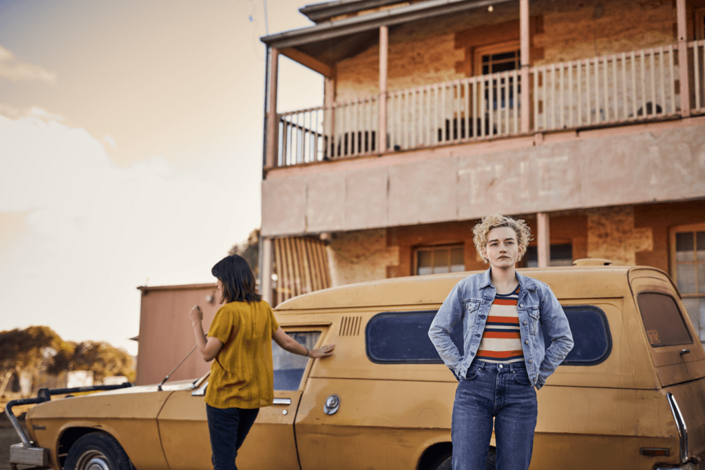 A film still from The Royal Hotel featuring a blond woman (Julia Garner) standing in front of a yellow car.