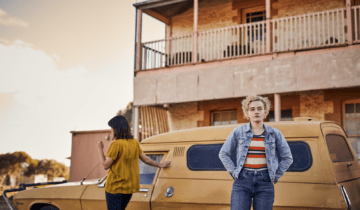 A film still from The Royal Hotel featuring a blond woman (Julia Garner) standing in front of a yellow car.