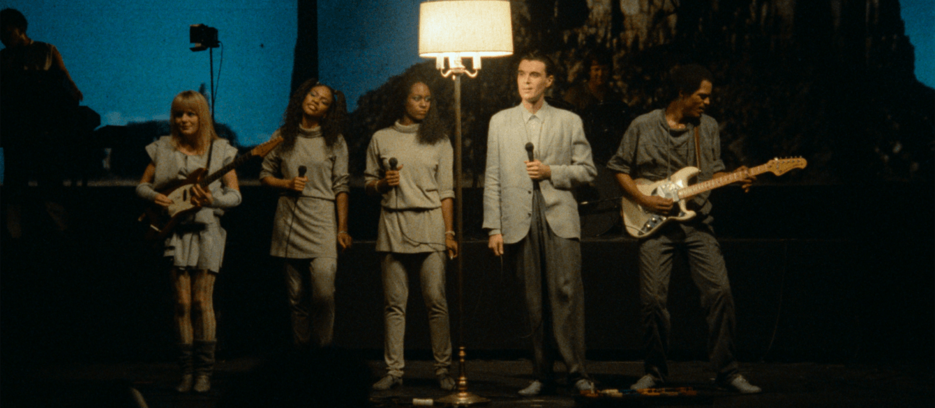 A film still from Stop Making Sense featuring David Byrne and The Talking Heads standing next to a lamp.