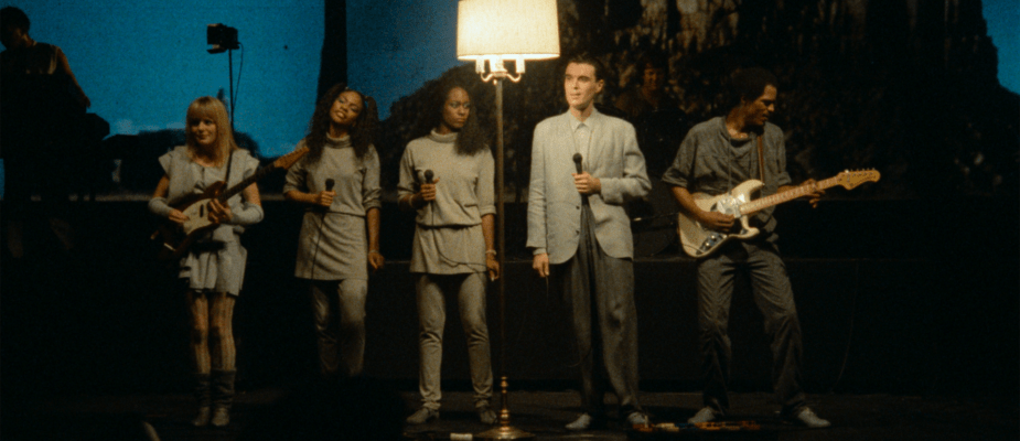 A film still from Stop Making Sense featuring David Byrne and The Talking Heads standing next to a lamp.
