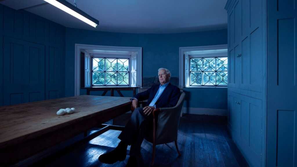 A still from The Pigeon Tunnel featuring a man ( spy novelist John le Carré) sitting in a chair next to a long table with eggs.
