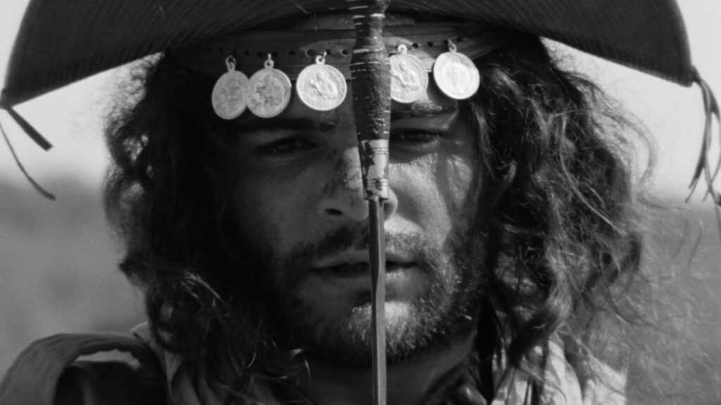 A film still from Black God, White Devil. A black-and-white image of a bearded man (Othon Bastos) wearing a headdress adorned with metal medallions stares straight ahead at the camera, his face divided in half by sword and its grip.