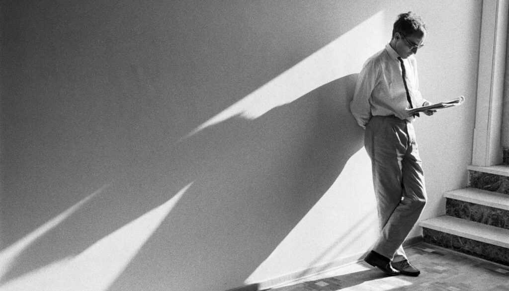 A film still from Godard Cinema. A black-and-white image of a man (Jean-Luc Godard) leaning against a wall reading, his long shadow cast out from him.