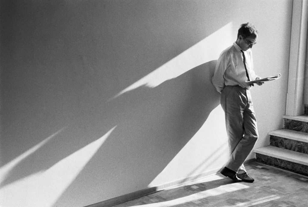 A film still from Godard Cinema. A black-and-white image of a man (Jean-Luc Godard) leaning against a wall reading, his long shadow cast out from him.