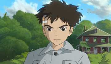 A film still from The Boy and the Heron. A boy with a bandage on the left side of his head stares straight ahead with a determined expression. Behind him are a two-story red house and a series of large trees behind it.