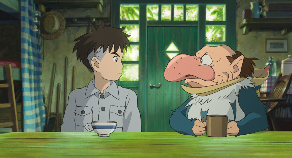 A film still from The Boy and the Heron. A boy and a squat, balding man with a large, beaked nose sit and look at each other tensely in a small cottage.