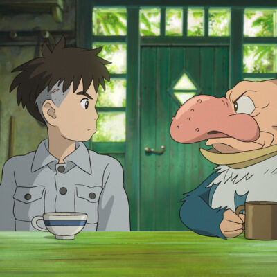 A film still from The Boy and the Heron. A boy and a squat, balding man with a large, beaked nose sit and look at each other tensely in a small cottage.