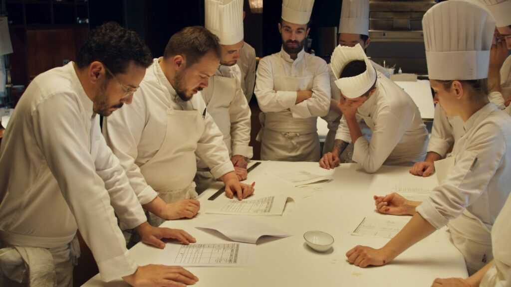 A Film Still from Menus-Plaisirs Les Troigros. A group of chefs stands around a long white counter poring over documents, mid-discussion.