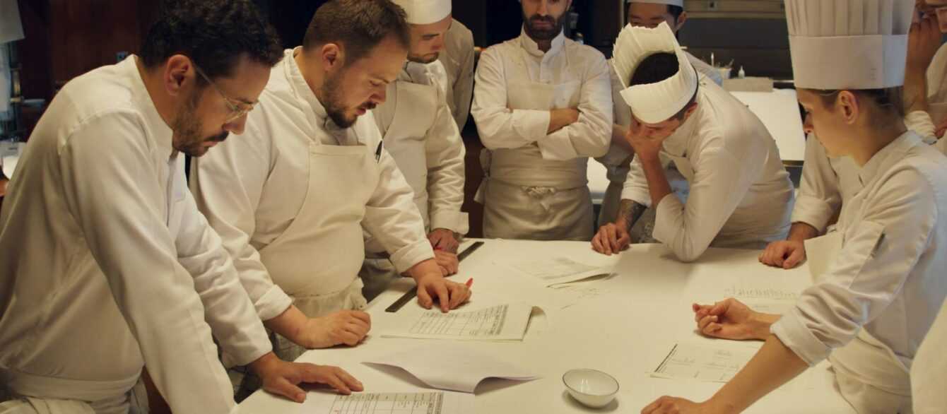 A Film Still from Menus-Plaisirs Les Troigros. A group of chefs stands around a long white counter poring over documents, mid-discussion.