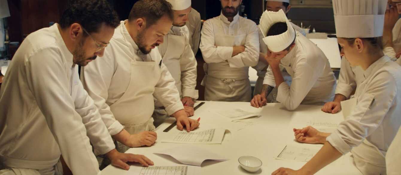 A Film Still from Menus-Plaisirs Les Troigros. A group of chefs stands around a long white counter poring over documents, mid-discussion.