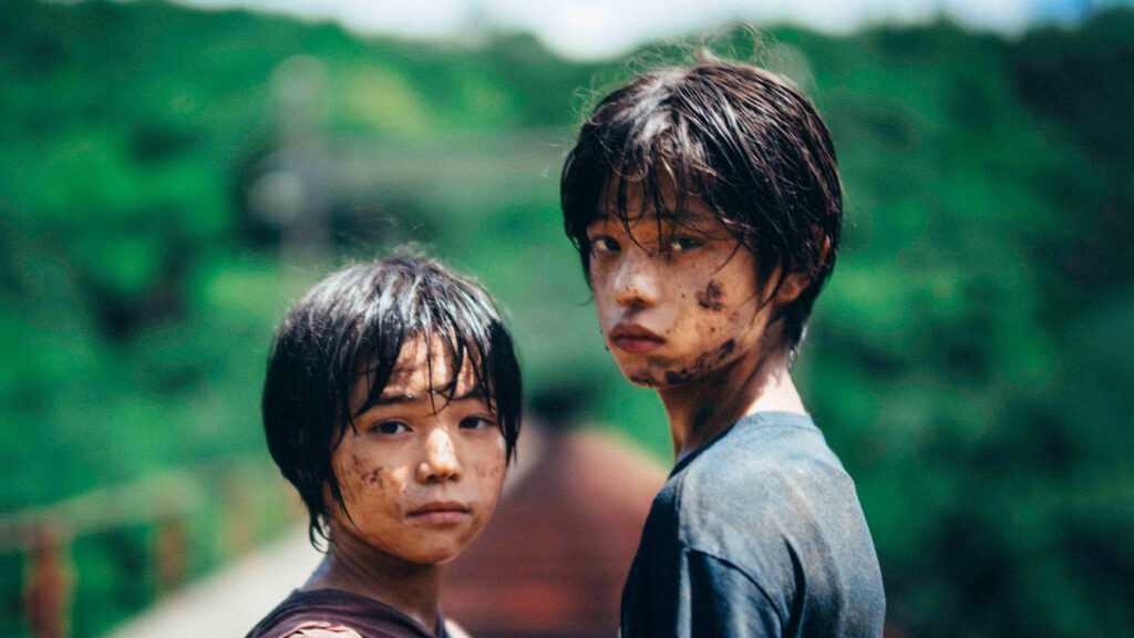 A film still from Monster. Two children (L: Hinata Hiiragi R: Soya Kurokawa) covered in dirt look back at the camera against a fuzzy, out-of-focus green hillside.