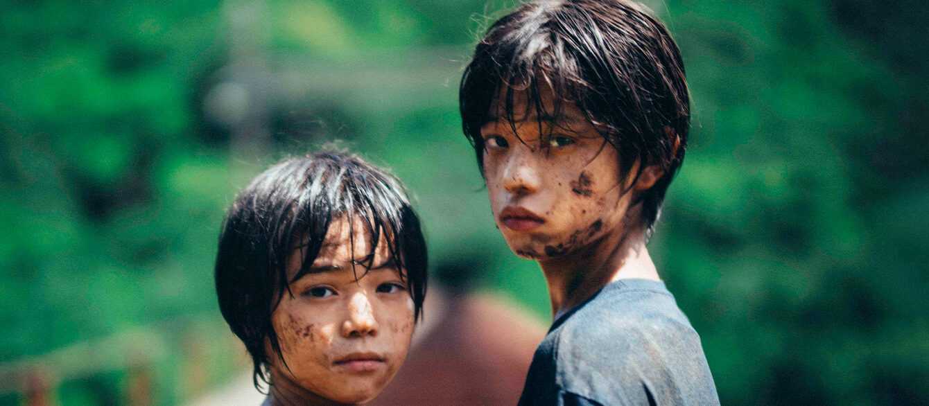 A film still from Monster. Two children (L: Hinata Hiiragi R: Soya Kurokawa) covered in dirt look back at the camera against a fuzzy, out-of-focus green hillside.