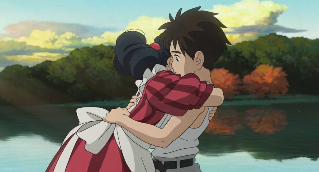 A film still from The Boy and the Heron. A boy and a girl wearing an elaborate stripped maroon dress hug in front of a placid lake with a forest in the distance.
