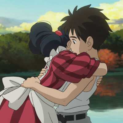 A film still from The Boy and the Heron. A boy and a girl wearing an elaborate stripped maroon dress hug in front of a placid lake with a forest in the distance.