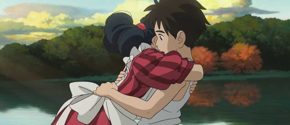 A film still from The Boy and the Heron. A boy and a girl wearing an elaborate stripped maroon dress hug in front of a placid lake with a forest in the distance.