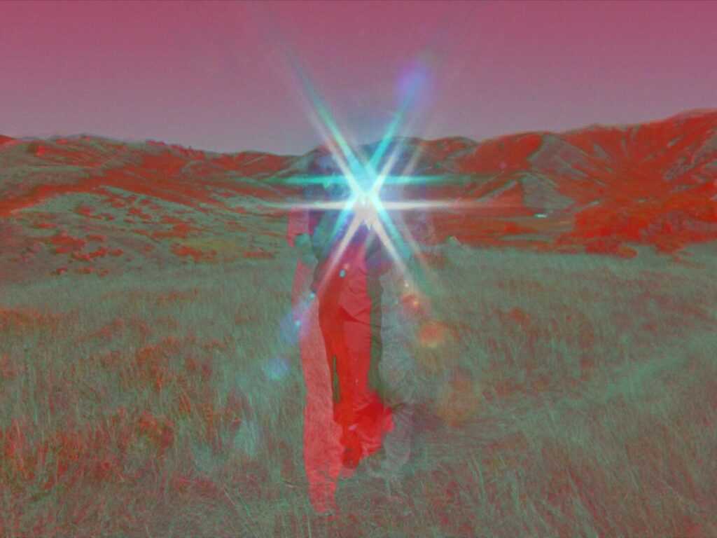 A Film Still from Last Things. A person stands in the middle of a grassy field surrounded by hills, the upper half of their body obscured by a star-shaped lens flare in the center of the image. The color of the image is affected by the camera’s filter; the person’s body and the hills behind them are a vibrant red, the sky a magenta, and the grass a mix of sage green and red.