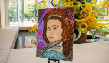 A painted portrait displayed on a small easel sits inside the museum with colorful glass artwork visible in the background.