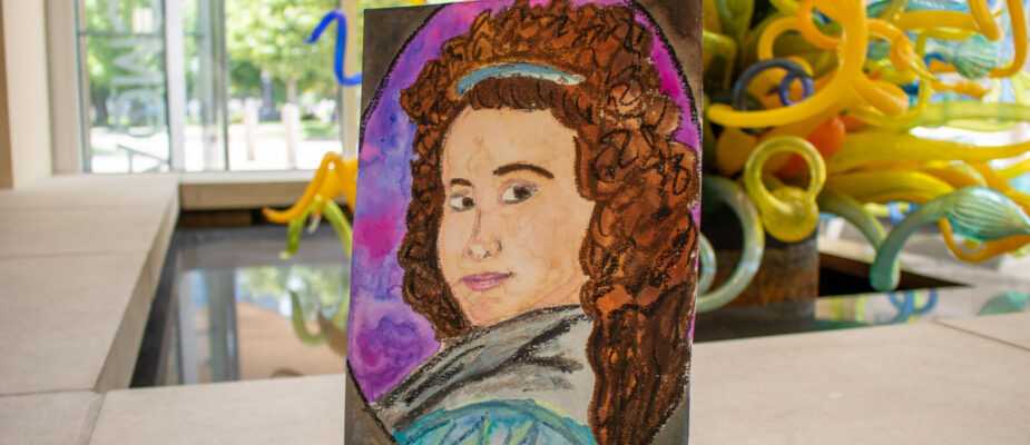 A painted portrait displayed on a small easel sits inside the museum with colorful glass artwork visible in the background.