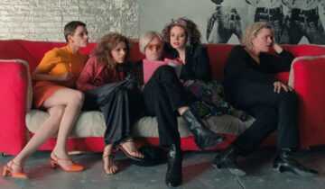 A group of people sit together on a red couch reviewing papers in a scene from the film I Shot Andy Warhol.