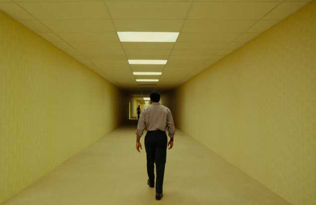 A man walks down a long, brightly lit corridor toward a distant figure in a scene from Backrooms.