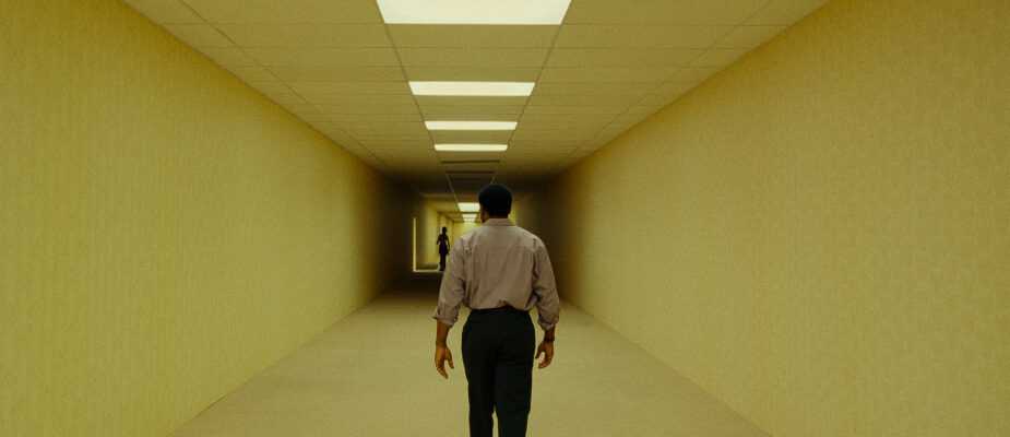 A man walks down a long, brightly lit corridor toward a distant figure in a scene from Backrooms.