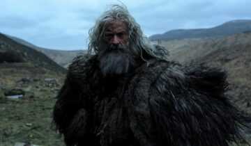 An older man with long gray hair and a fur cloak stands in a rugged mountain landscape in a scene from The Death of Robin Hood.