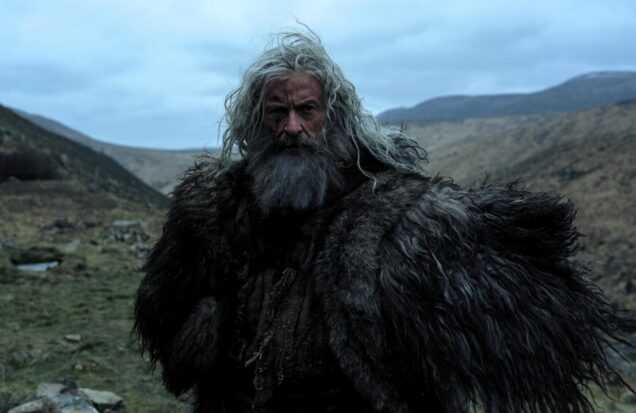 An older man with long gray hair and a fur cloak stands in a rugged mountain landscape in a scene from The Death of Robin Hood.