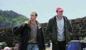 Two young men walk beside stone walls and fishing crates in a coastal setting in a scene from Rose of Nevada.