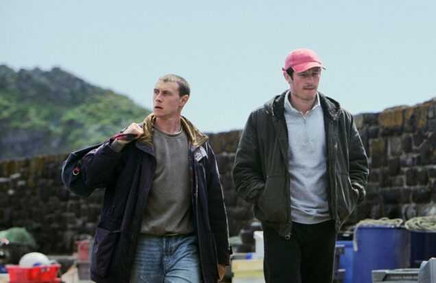 Two young men walk beside stone walls and fishing crates in a coastal setting in a scene from Rose of Nevada.