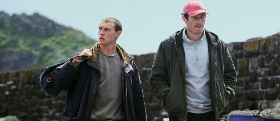 Two young men walk beside stone walls and fishing crates in a coastal setting in a scene from Rose of Nevada.
