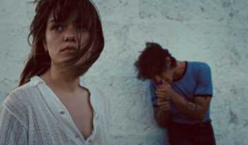 A woman in a white blouse stands in the foreground while a man stands against a wall in a dramatic scene from Romeria.