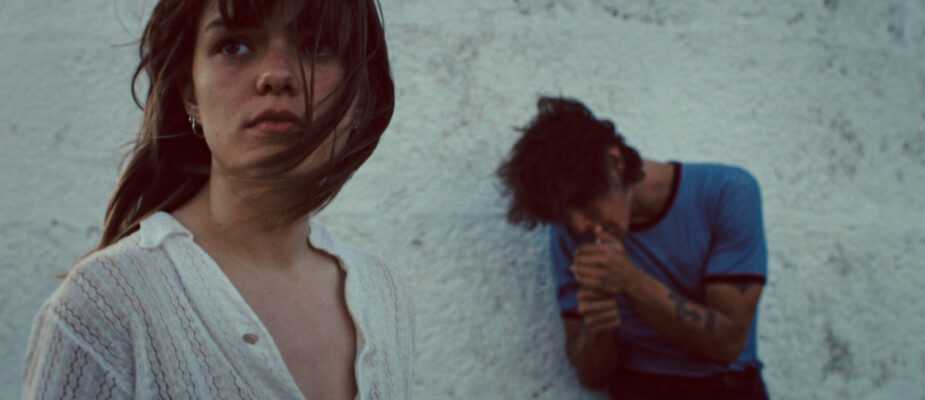 A woman in a white blouse stands in the foreground while a man stands against a wall in a dramatic scene from Romeria.