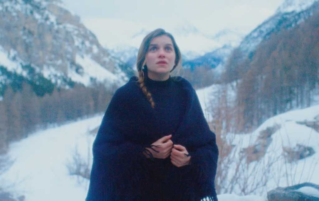 A woman wrapped in a dark shawl stands in a snowy mountain valley in a scene from The Girl in the Snow.