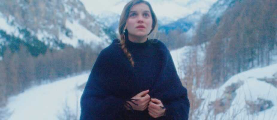 A woman wrapped in a dark shawl stands in a snowy mountain valley in a scene from The Girl in the Snow.