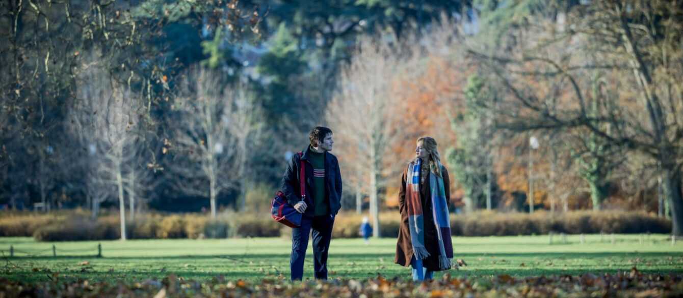 Two people walk through a park covered in autumn leaves while talking in a scene from Two Pianos.