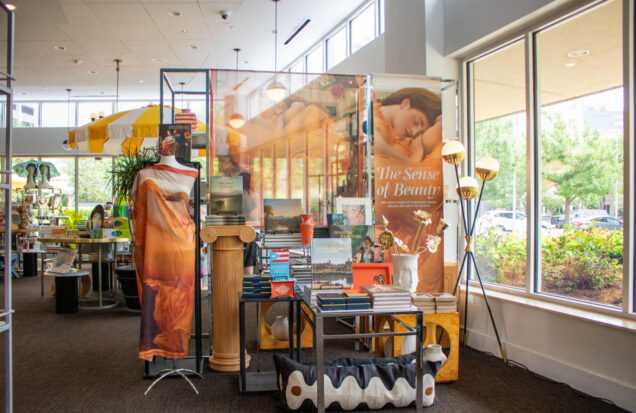 Museum store display featuring The Sense of Beauty merchandise, books, textiles, and decorative displays near large windows.